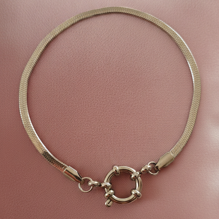 Primer plano de una pulsera plateada de cadena plana y lisa (Herringbone o serpiente) con un cierre de aro redondo y prominente (tipo toggle) sobre una tela de terciopelo rosa pastel. Joyería de moda minimalista en acero inoxidable color plata.