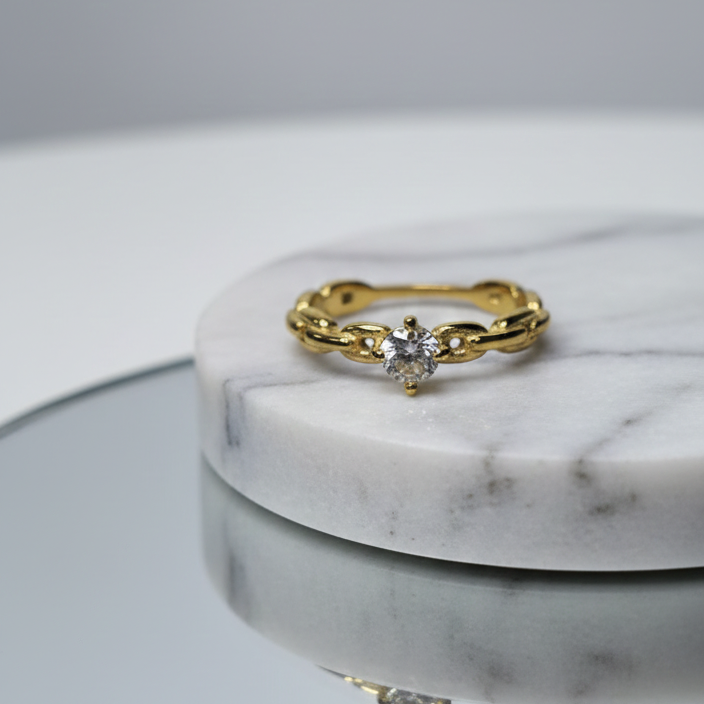 Anillo solitario dorado con una zirconia central en un engaste retorcido o trenzado, sobre una superficie de espejo. Anillo de compromiso o promesa en acero inoxidable baño de oro 18k. Joyería elegante femenina de nudo.